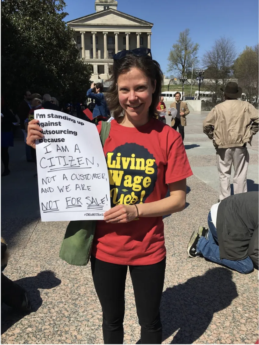 A photo of Sarah Vandergrift Eldridge holding a sign that reads "I'm standing up against outsourcing because I am a citizen, not a customer and we are not for sale!"